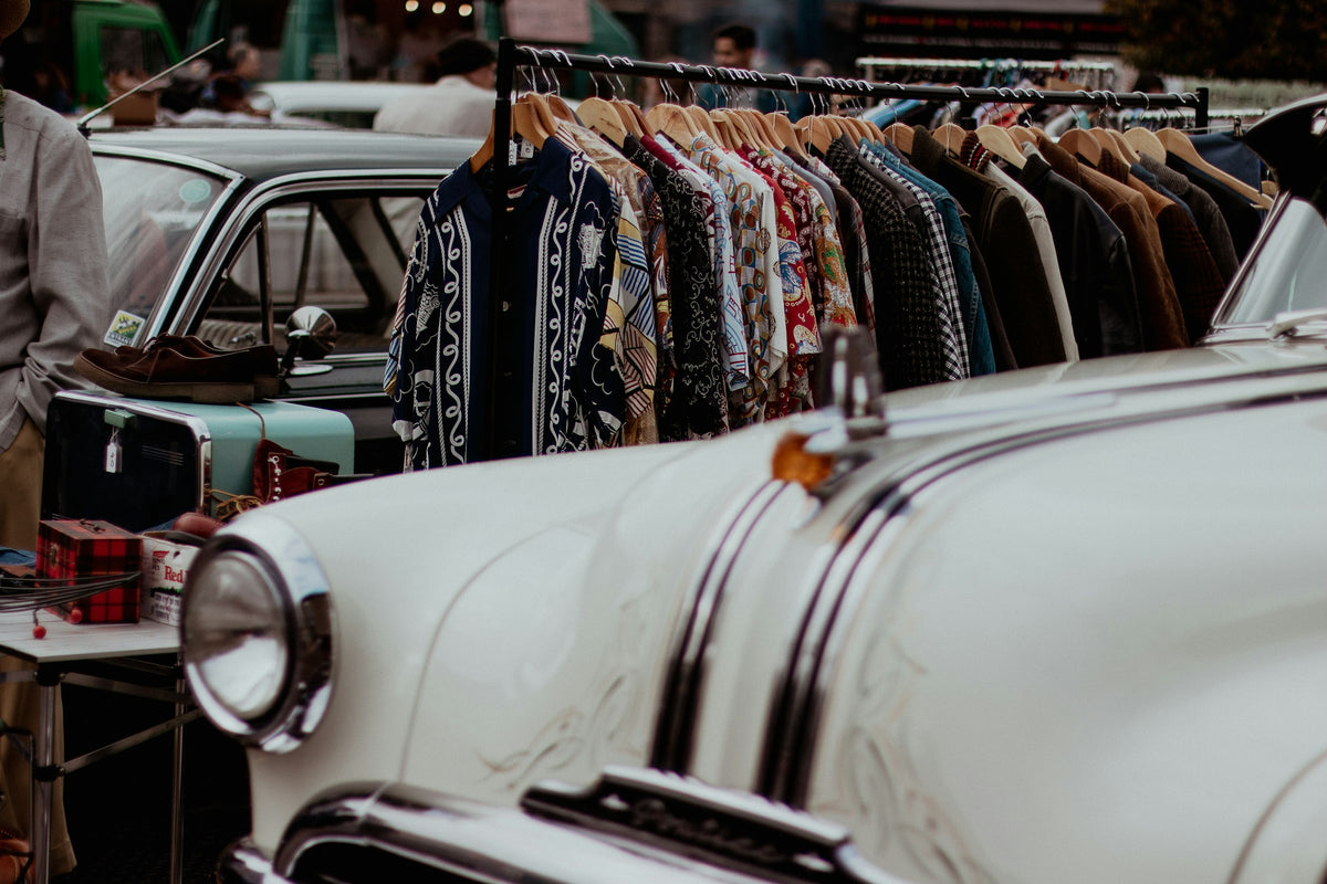 A rail of second-hand shirts hung outdoors beside a parked car, seen in flat daylight.