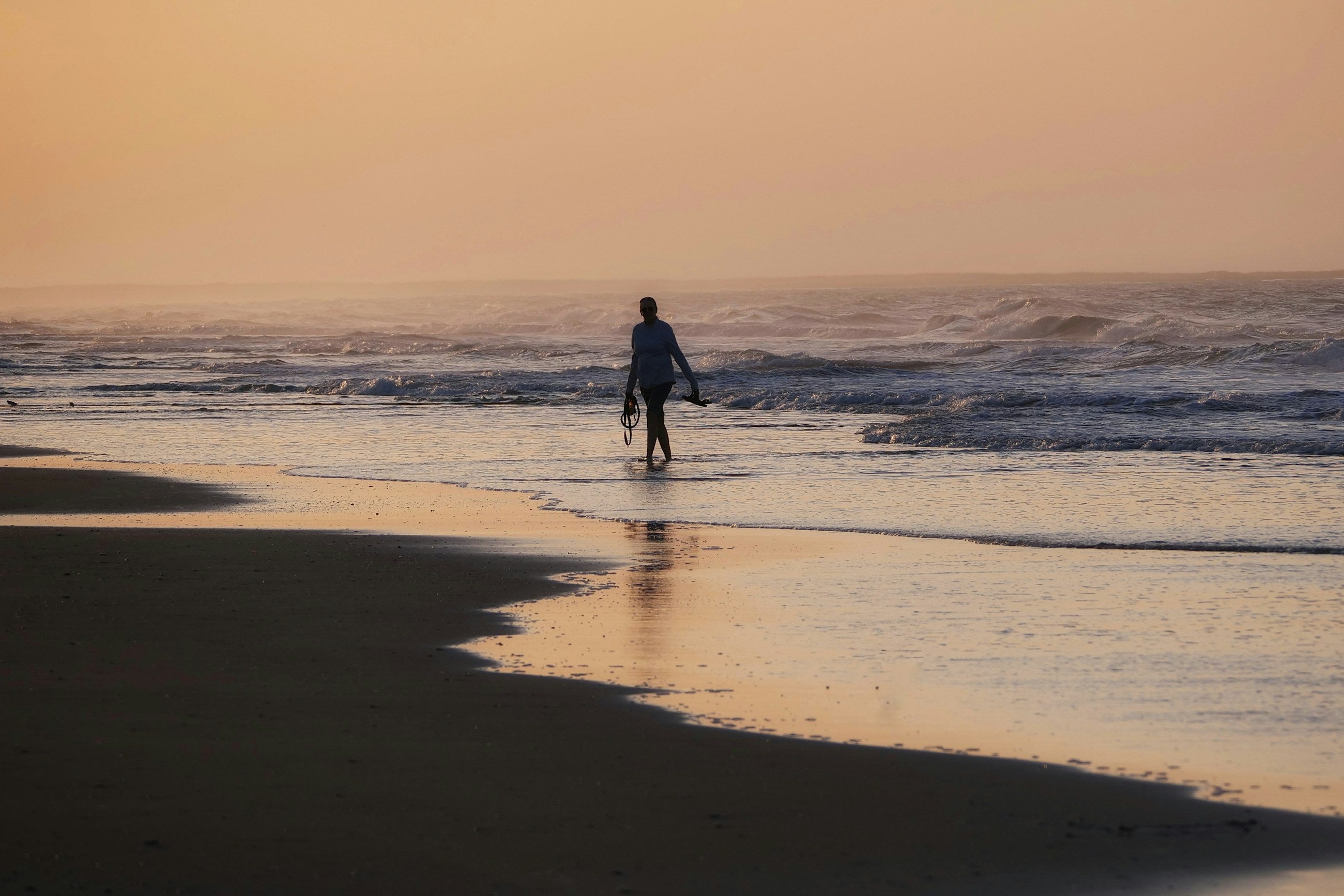 A lone surfer walking out into the shallows on an empty beach, board under one arm.