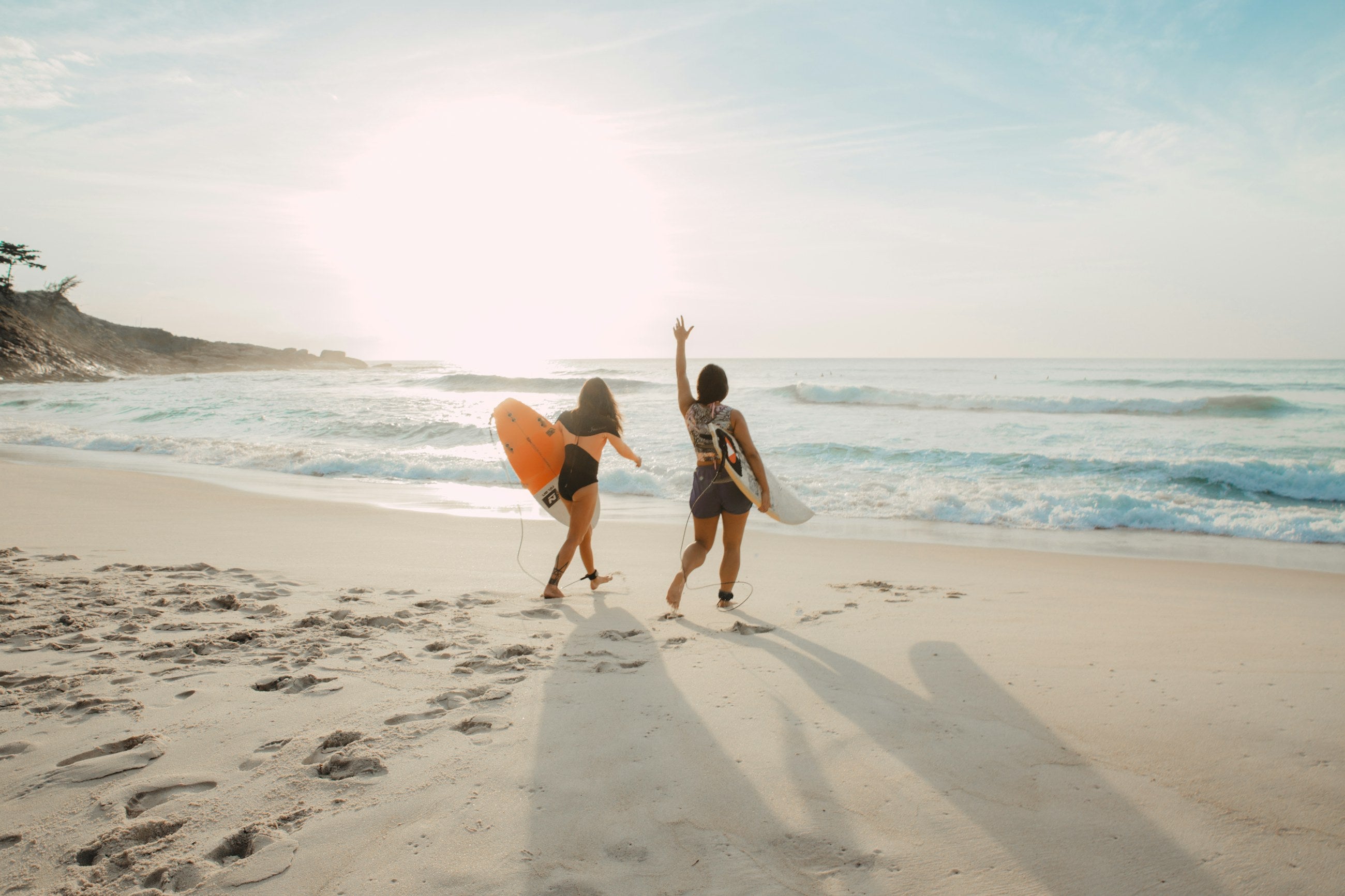 Two surfers walking down to the water with their boards, one with an arm raised, on wide pale sand.