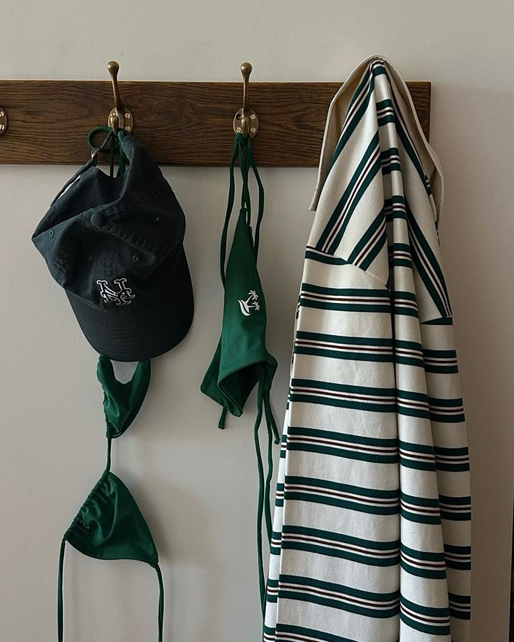 A cap, green swimwear and a striped towel hanging from brass hooks on a wooden peg rail.