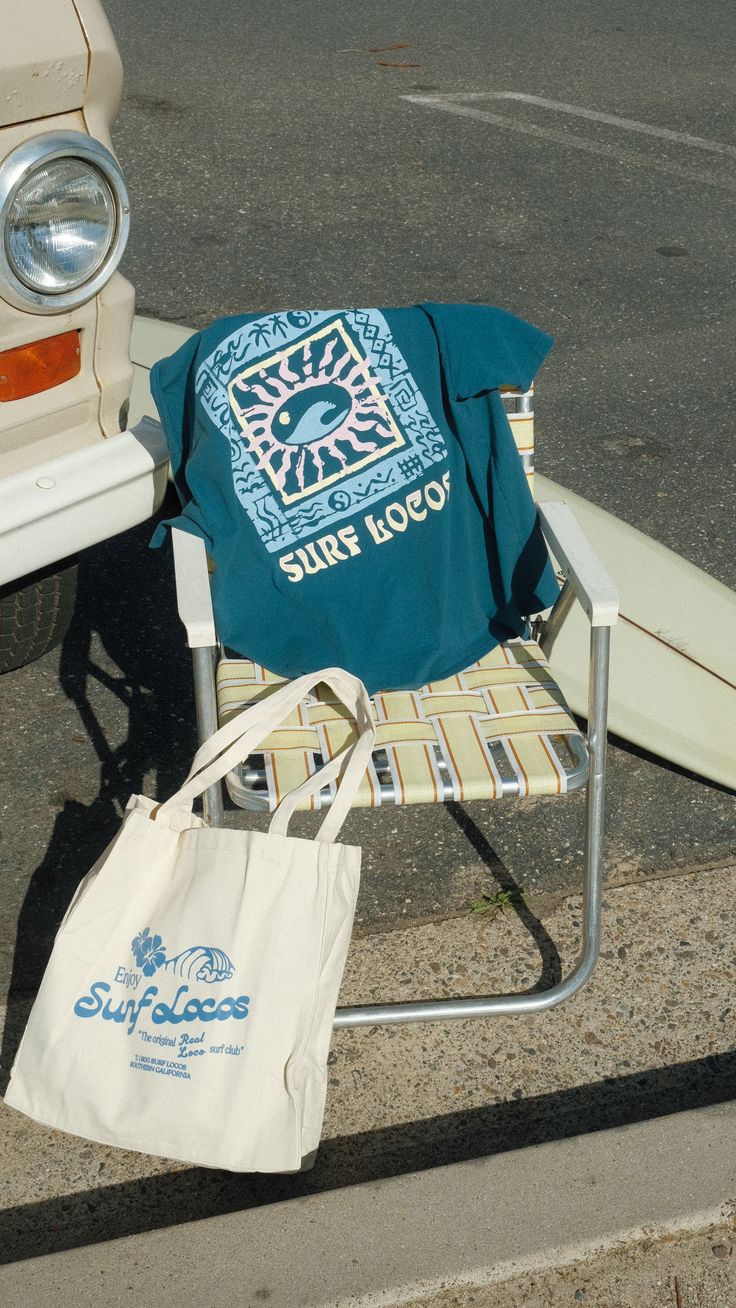 A vintage surf t-shirt over a folding beach chair, tote bag and surfboard beside a cream-coloured car.